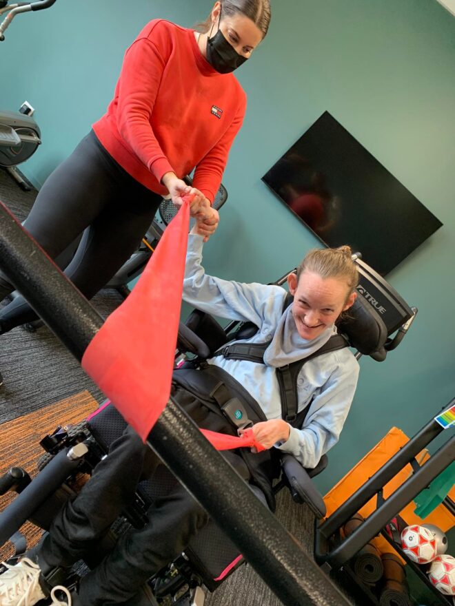 Girl in wheelchair smiling at camera while doing arm exercises with ribbon
